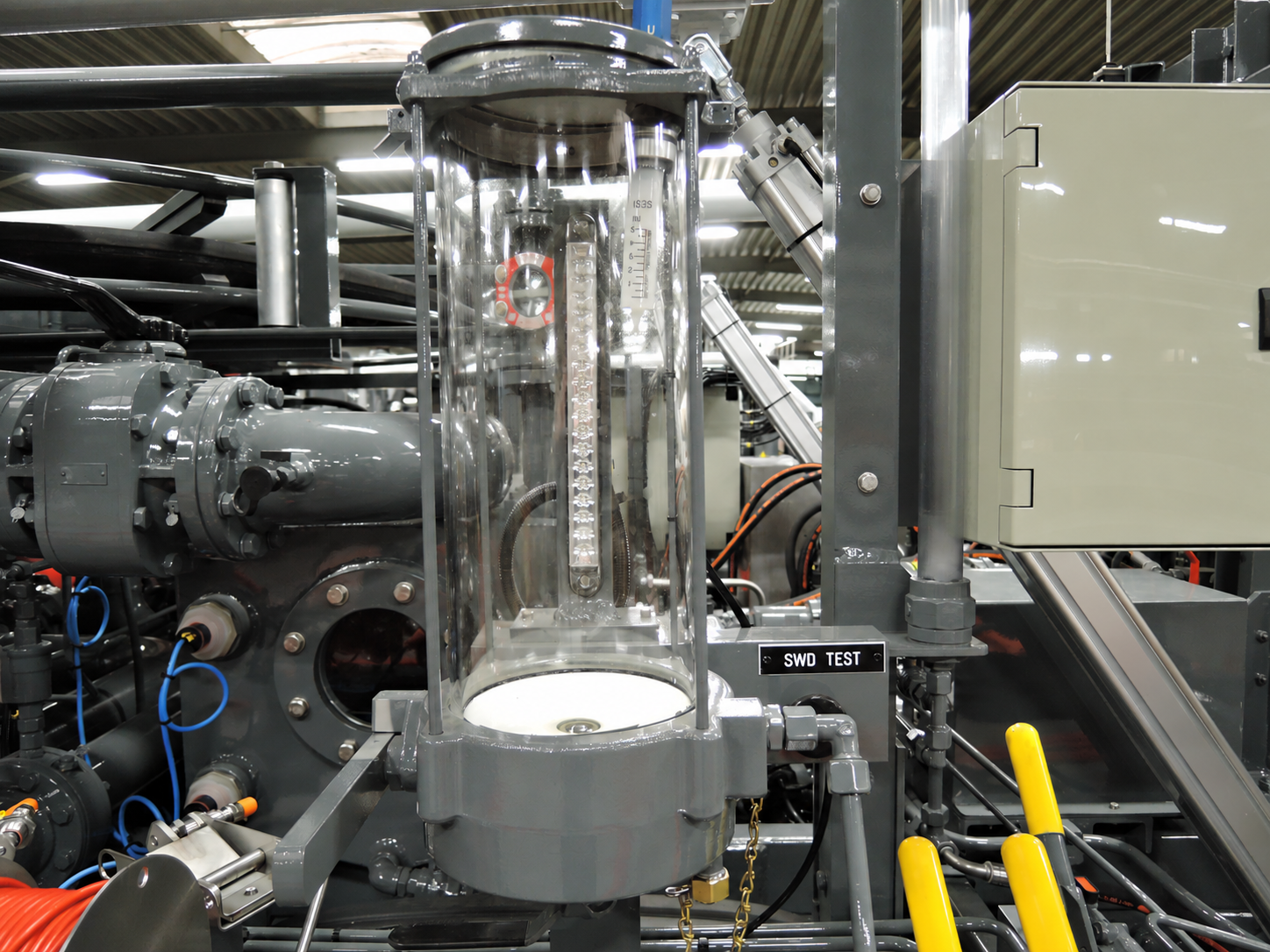 Transparent cylindrical sampling chamber mounted beside grey pipework, with an SWD TEST label.