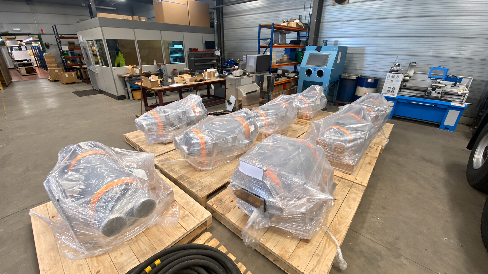 Six metal component assemblies wrapped in clear plastic and arranged on wooden pallets inside a workshop.